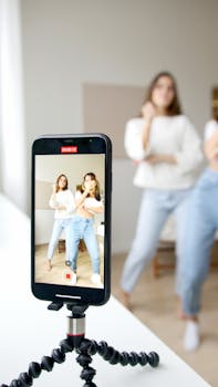 Two young women dancing in front of a smartphone camera on a tripod indoors.