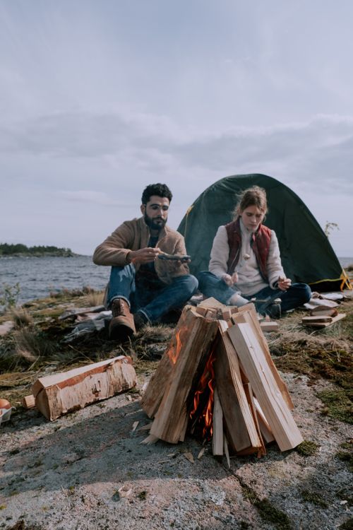 Free Couple enjoying a peaceful summer campfire by the river with a green tent nearby, surrounded by nature. Stock Photo