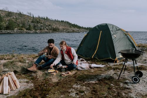 Free Couple enjoying a picnic by a tent and grill near a serene riverside camping site. Stock Photo