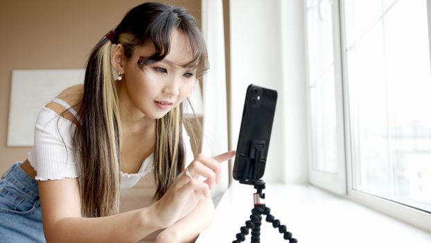Asian woman using smartphone with a tripod by a window in a bright indoor setting.