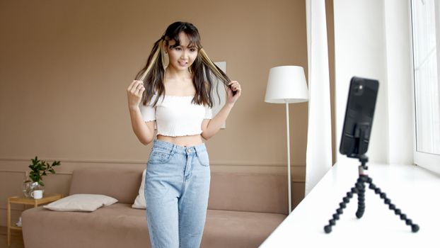 Asian woman in denim jeans and white top vlogging with smartphone on tripod in a modern living room.