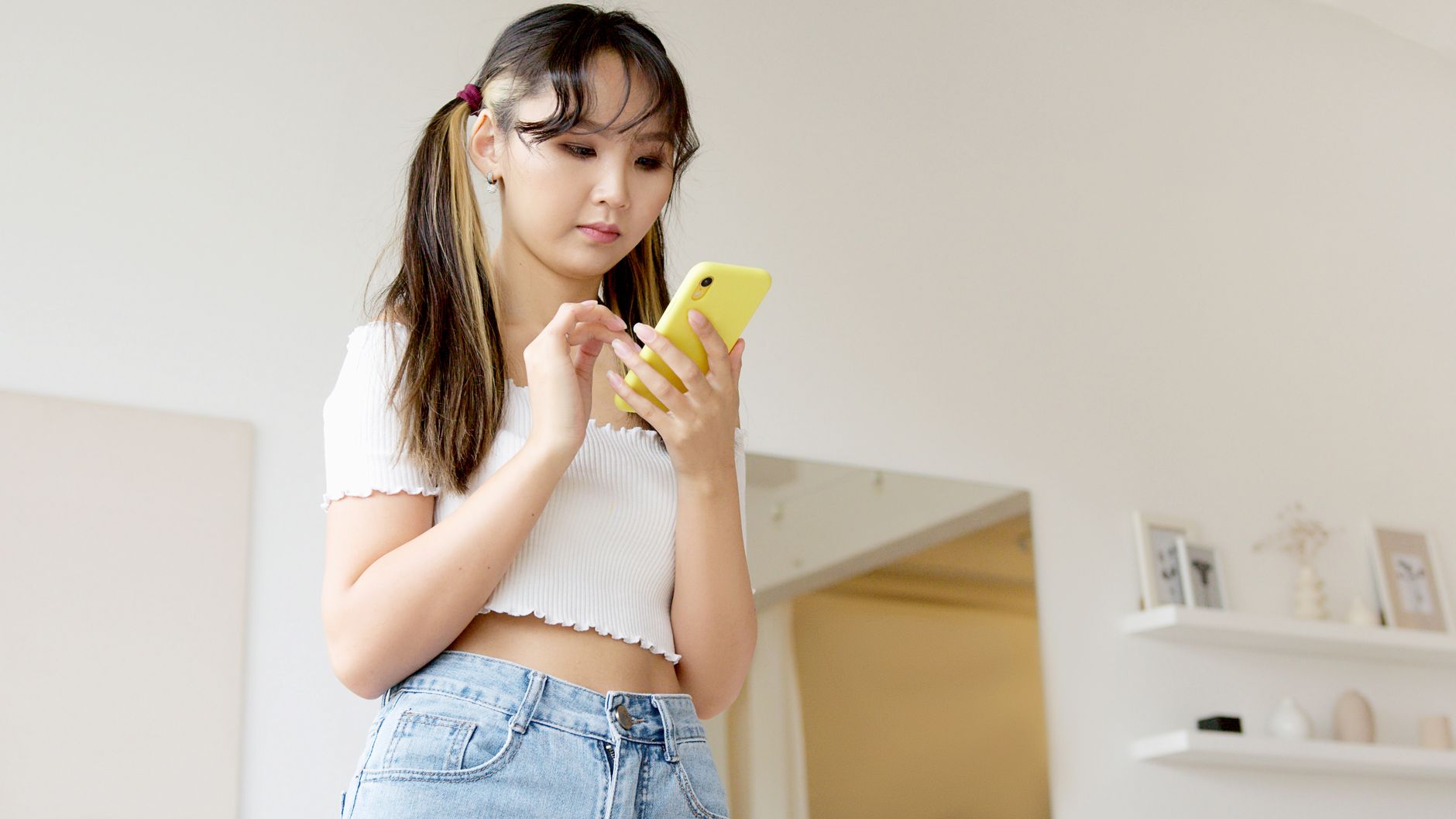 A young Asian woman using a smartphone inside a modern home setting, focusing intently on the screen.