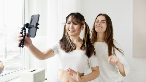 Free Two women joyfully taking a selfie indoors with a smartphone and tripod, capturing a fun moment. Stock Photo
