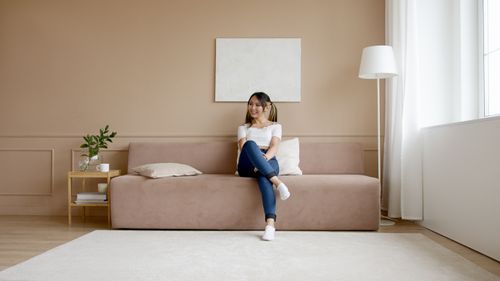 Free A smiling woman sits comfortably on a minimalist beige sofa in a stylish modern living room. Stock Photo