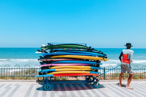 Free Back view full body of anonymous barefoot male in hat pulling cart with surfboards while walking on seafront with fence near waving ocean Stock Photo