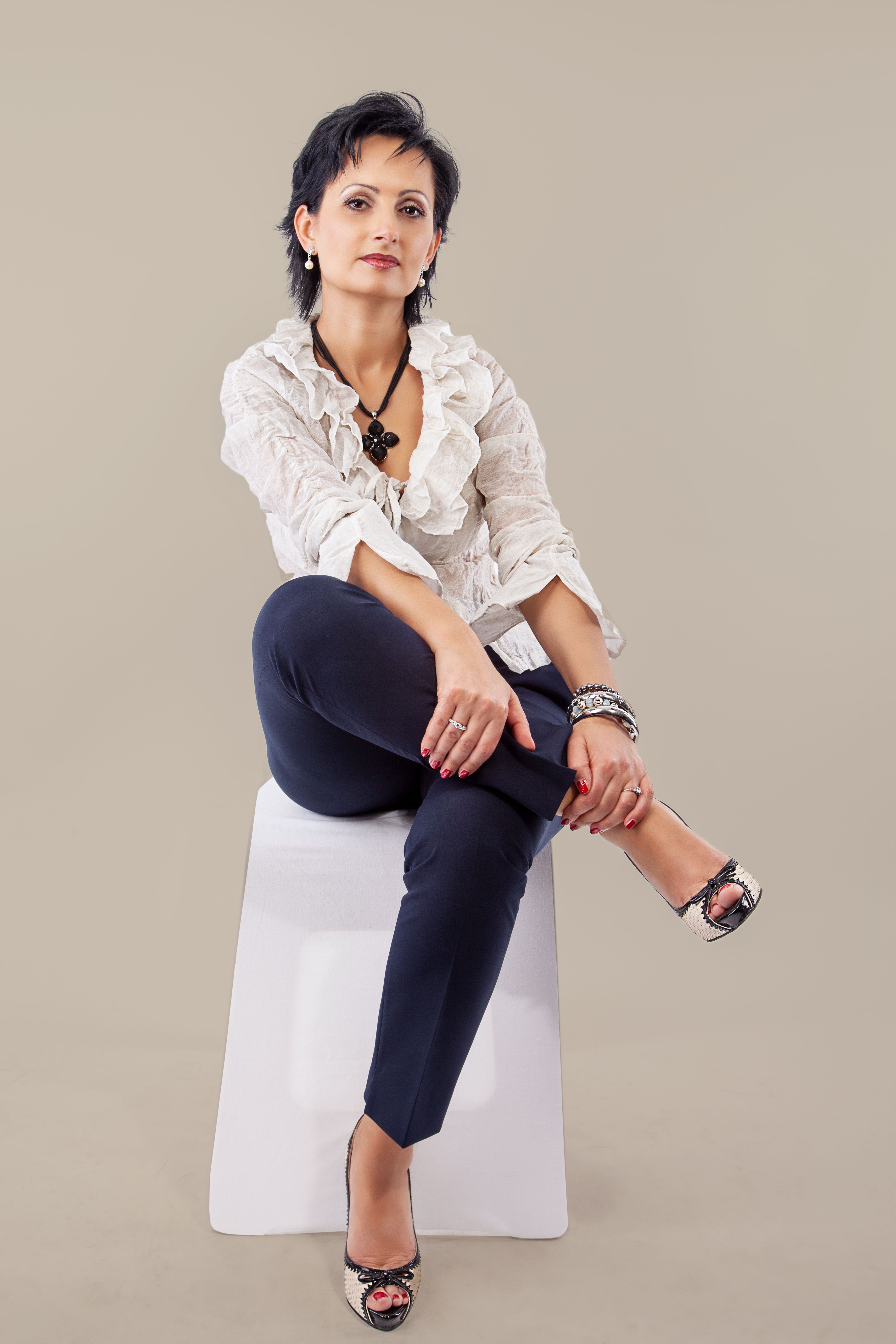 Stylish woman in white blouse and accessories posing on stool in studio.