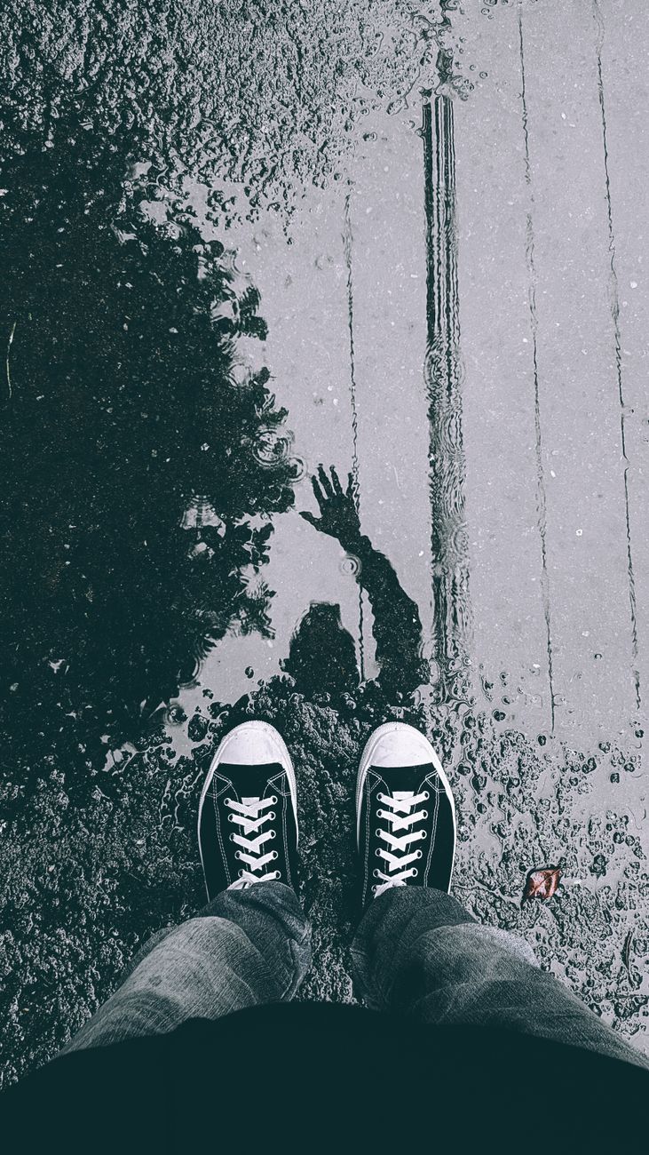 Grayscale image of sneakers by a puddle, reflecting a waving silhouette on a wet street.