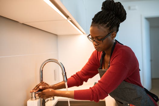 An adult woman with dreadlocks washing hands at a kitchen sink, emphasizing hygiene and cleanliness.