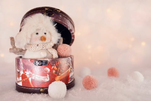 Small decorative snowman in hat and scarf placed in round box on white surface with artificial snowballs during Christmas holiday