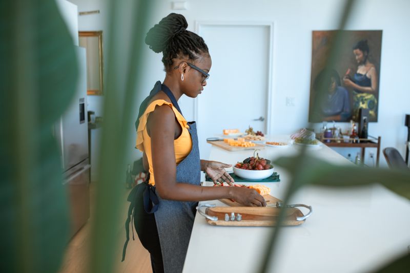 A Black woman in an apron preparing fresh produce on a cutting board at a bright kitchen counter at home.