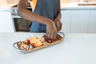 Person arranging a charcuterie board with nuts, cheese, and fruits on a wooden platter.