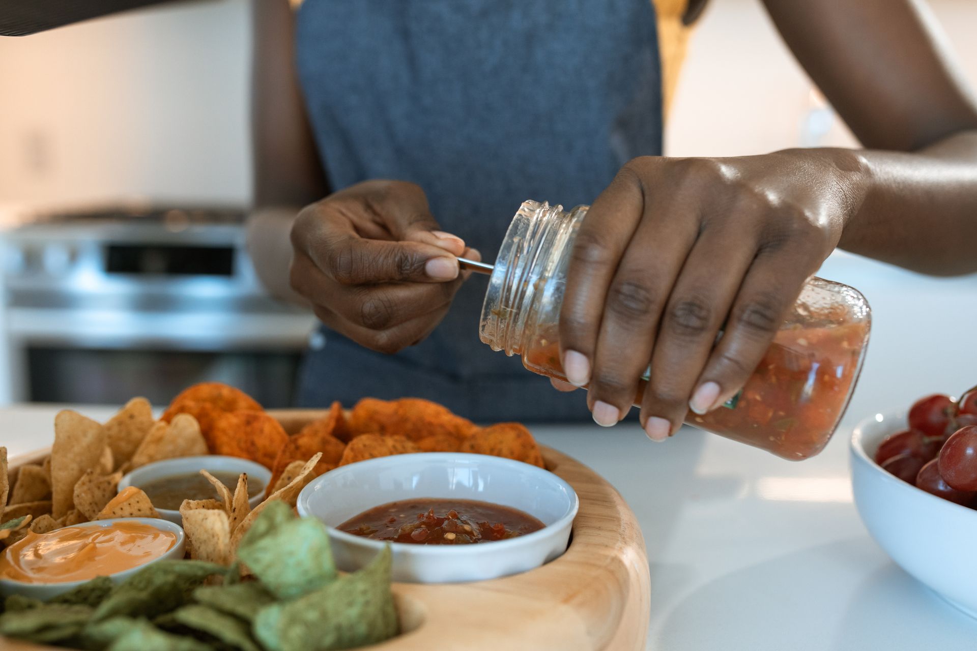 A person pouring salsa onto a snack platter with tortilla chips and grapes.