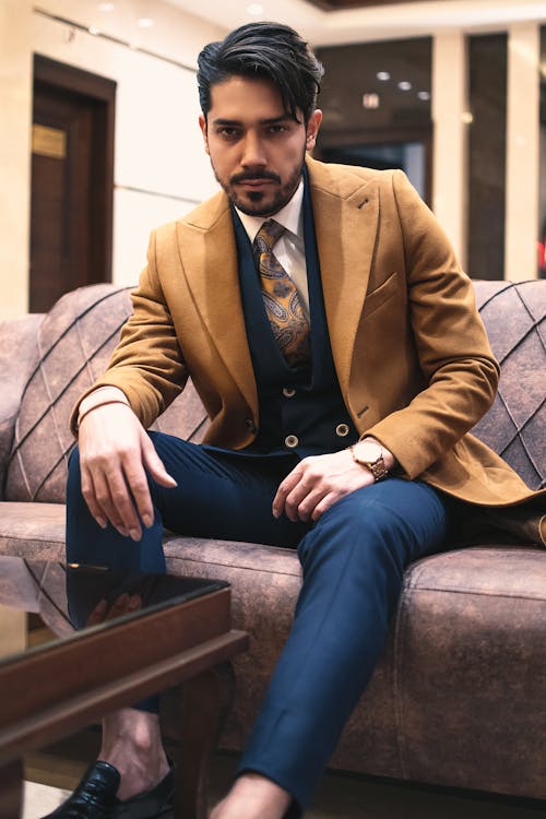 Confident adult ethnic bearded businessman wearing stylish colorful suit sitting on comfortable couch in business center and looking at camera