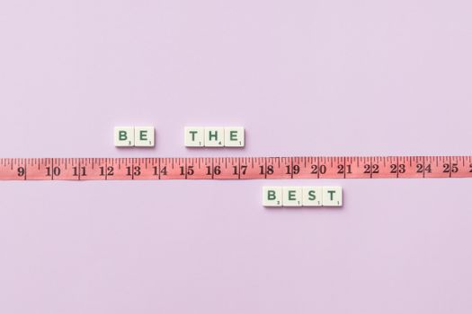 Scrabble tiles spell 'Be the Best' next to a measuring tape on a pastel background.