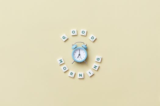 Aesthetic flat lay with a blue alarm clock surrounded by scrabble tiles spelling 'Good Morning' on a pastel background.
