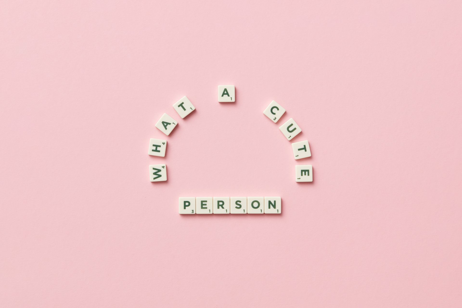 Flatlay of Scrabble tiles spelling 'What a cute person' on a pastel pink background.
