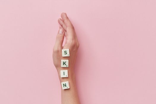 A creative arrangement of Scrabble tiles spelling 'skin' on a hand against a pink backdrop.
