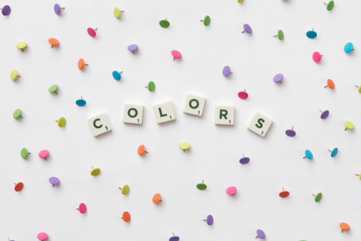 Vibrant multicolored thumbtacks and 'colors' spelled with Scrabble tiles on a white background.