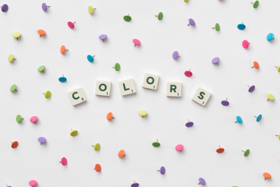 Vibrant multicolored thumbtacks and 'colors' spelled with Scrabble tiles on a white background.