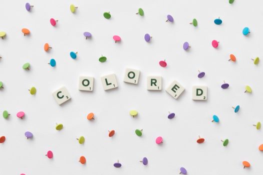 Bright multicolored thumbtacks with scrabble tiles spelling 'COLORED' on a white backdrop.