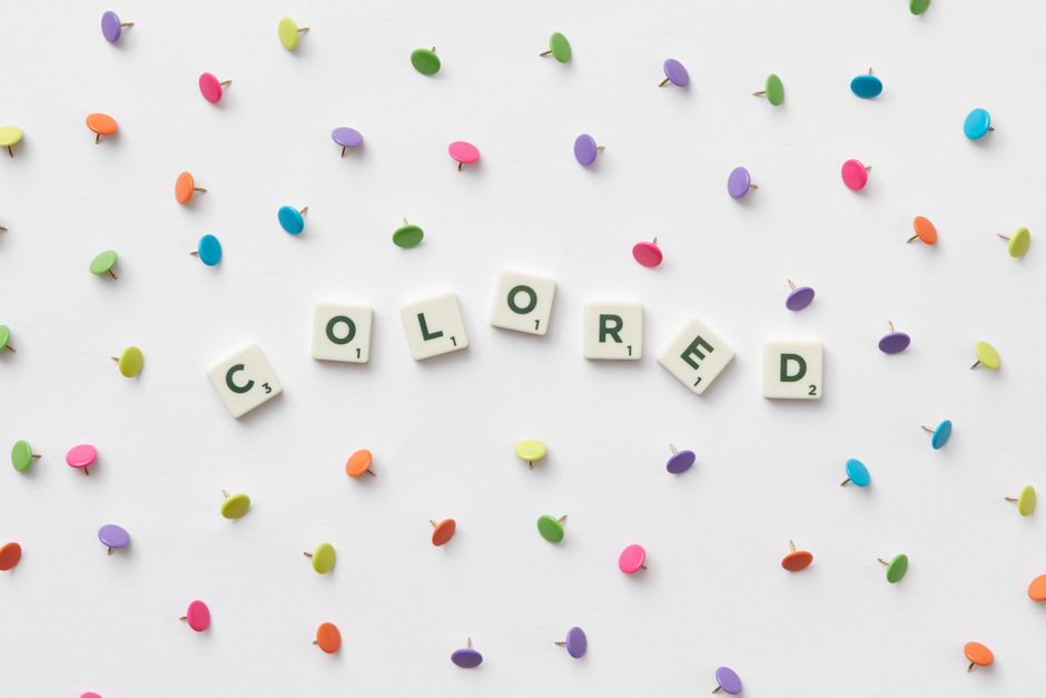 Bright multicolored thumbtacks with scrabble tiles spelling 'COLORED' on a white backdrop.