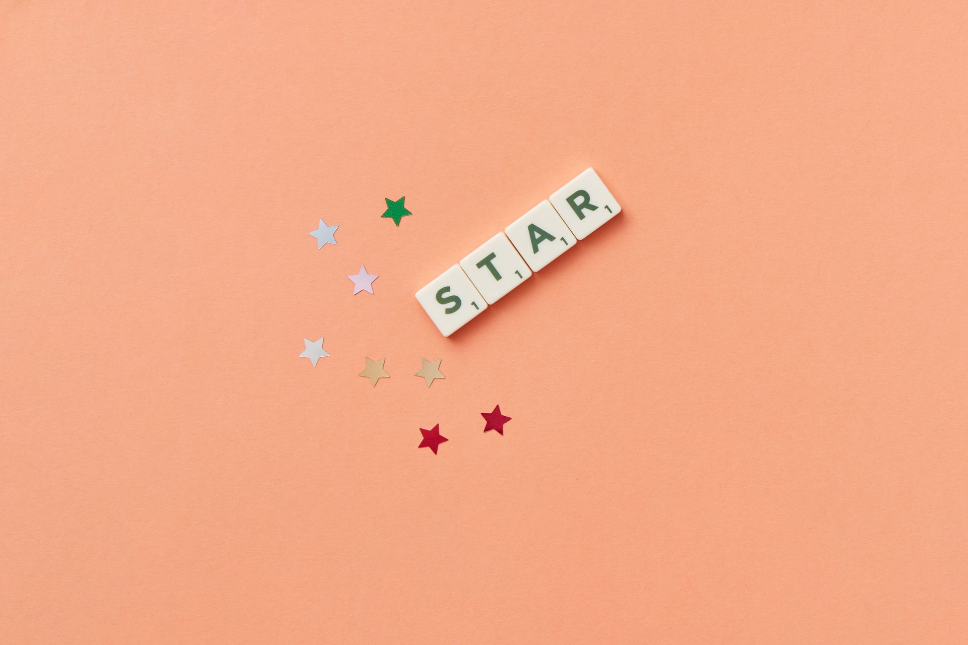 Flat lay image of Scrabble letters spelling 'STAR' on a peach background with colorful stars.