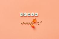 A minimalist composition featuring Scrabble tiles spelling 'woman' with delicate orange flowers on a peach background.