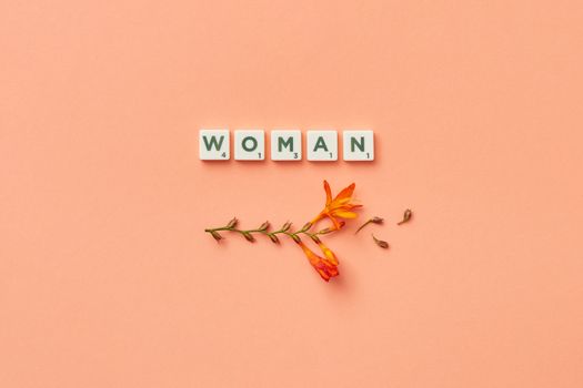 A minimalist composition featuring Scrabble tiles spelling 'woman' with delicate orange flowers on a peach background.