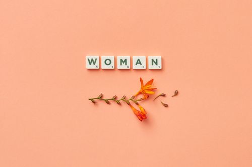 Free A minimalist composition featuring Scrabble tiles spelling 'woman' with delicate orange flowers on a peach background. Stock Photo