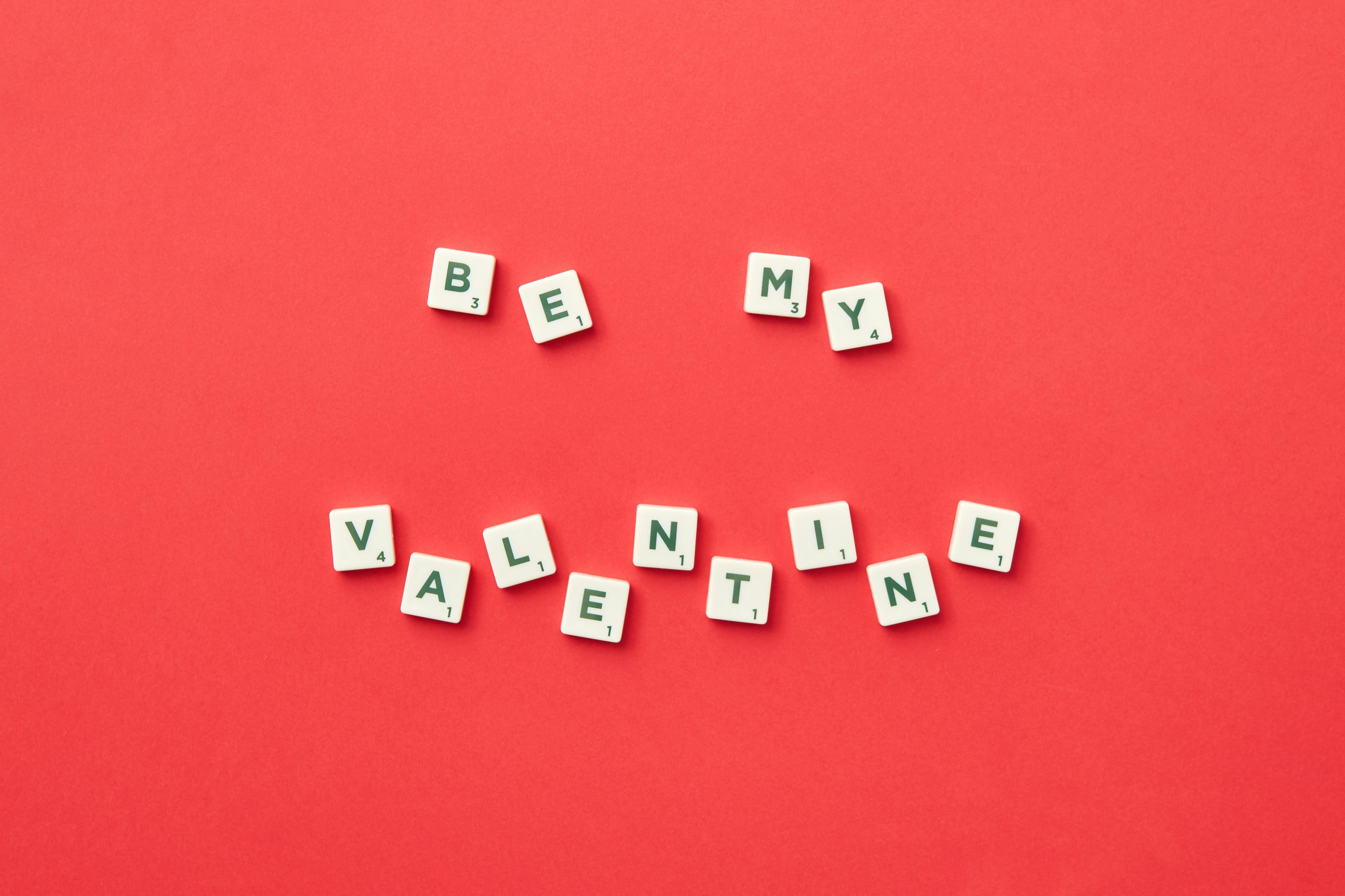 Scrabble tiles spell 'Be My Valentine' on a red background, perfect for Valentine's Day.