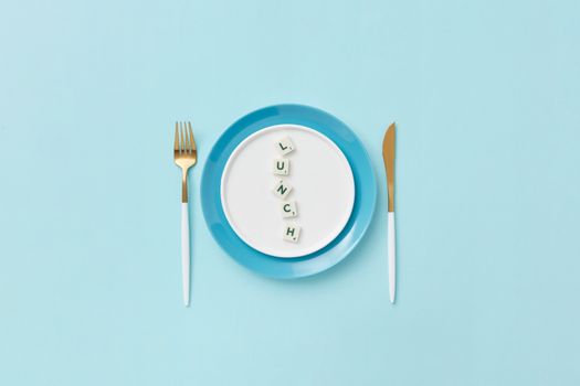 Flat lay of a lunch setting with Scrabble letters spelling 'lunch' on a blue plate and cutlery.