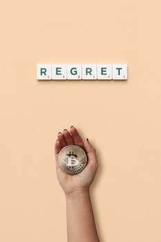 A hand holding a Bitcoin under 'regret' spelled with tiles on a beige background.