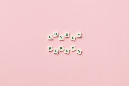Free Scrabble tiles spell 'lovely person' on a pastel pink background, promoting positivity. Stock Photo