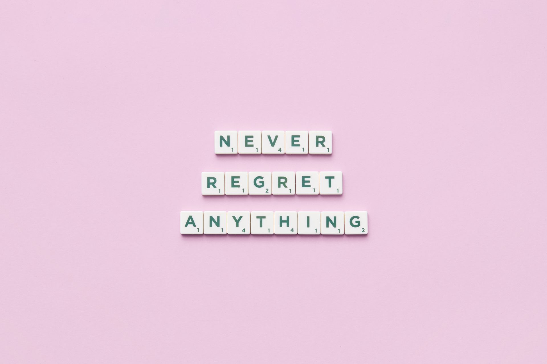 Scrabble tiles spelling 'Never Regret Anything' on a pink background in a flat lay style.