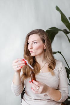 A woman with long hair uses a dropper with a brown bottle indoors, by plants.