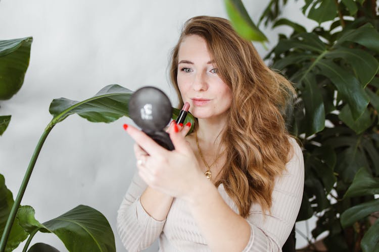 Photo Of A Woman With Blond Hair Putting On Pink Lipstick Near Green Plants