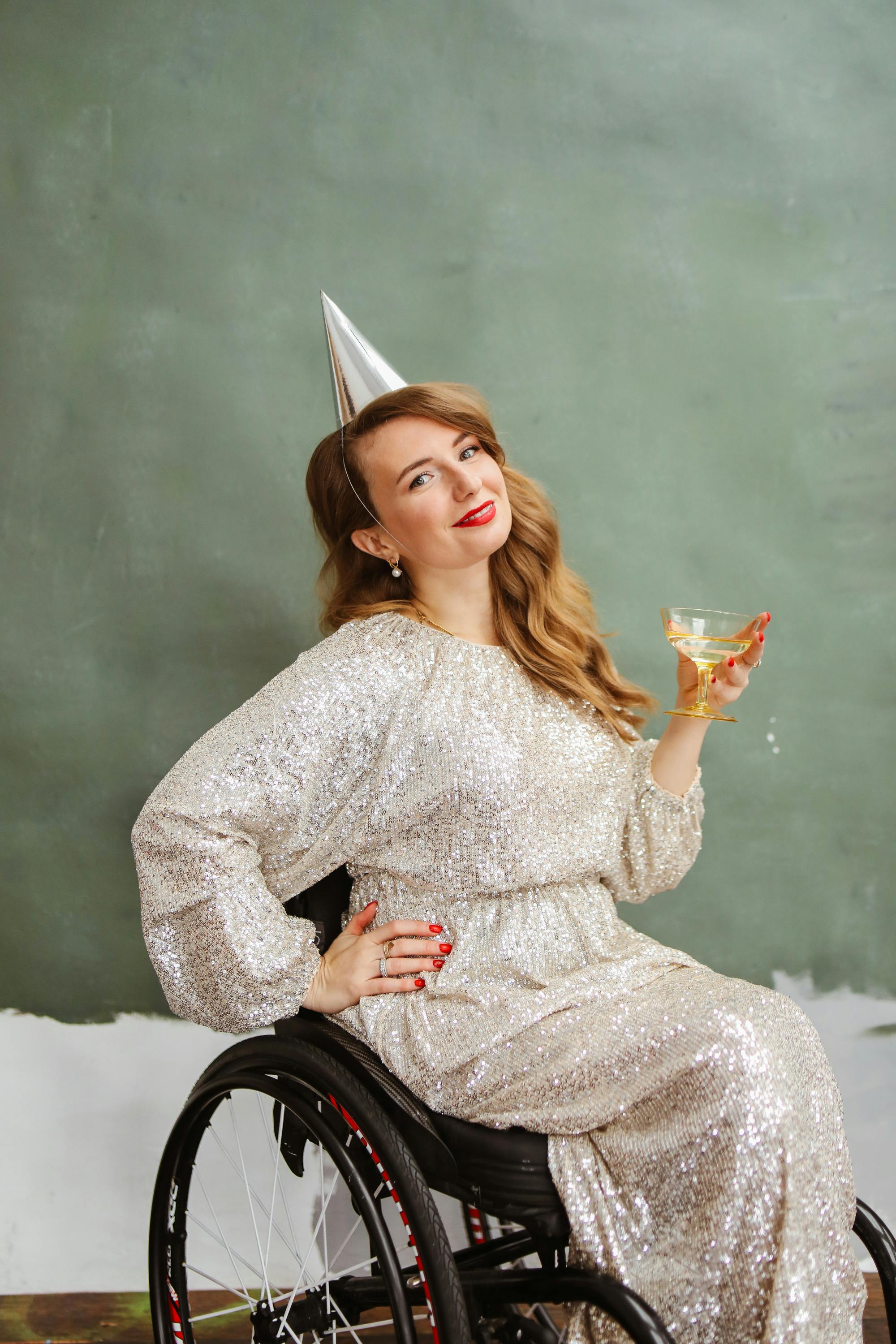 Elegant woman in a silver dress and party hat celebrating with a cocktail in a wheelchair.