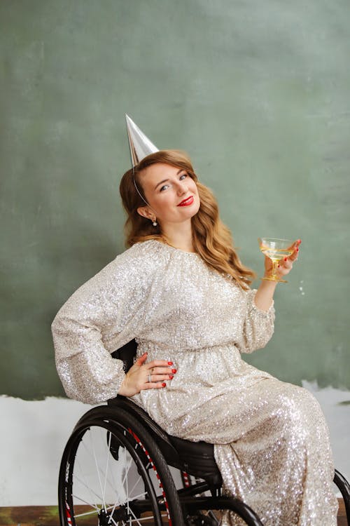 A Woman in Silver Dress Sitting on the Wheelchair while Holding a Cocktail Glass