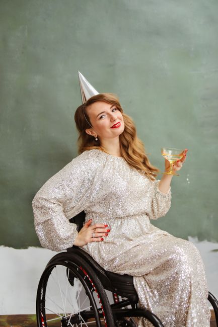 Elegant woman in a silver dress and party hat celebrating with a cocktail in a wheelchair.