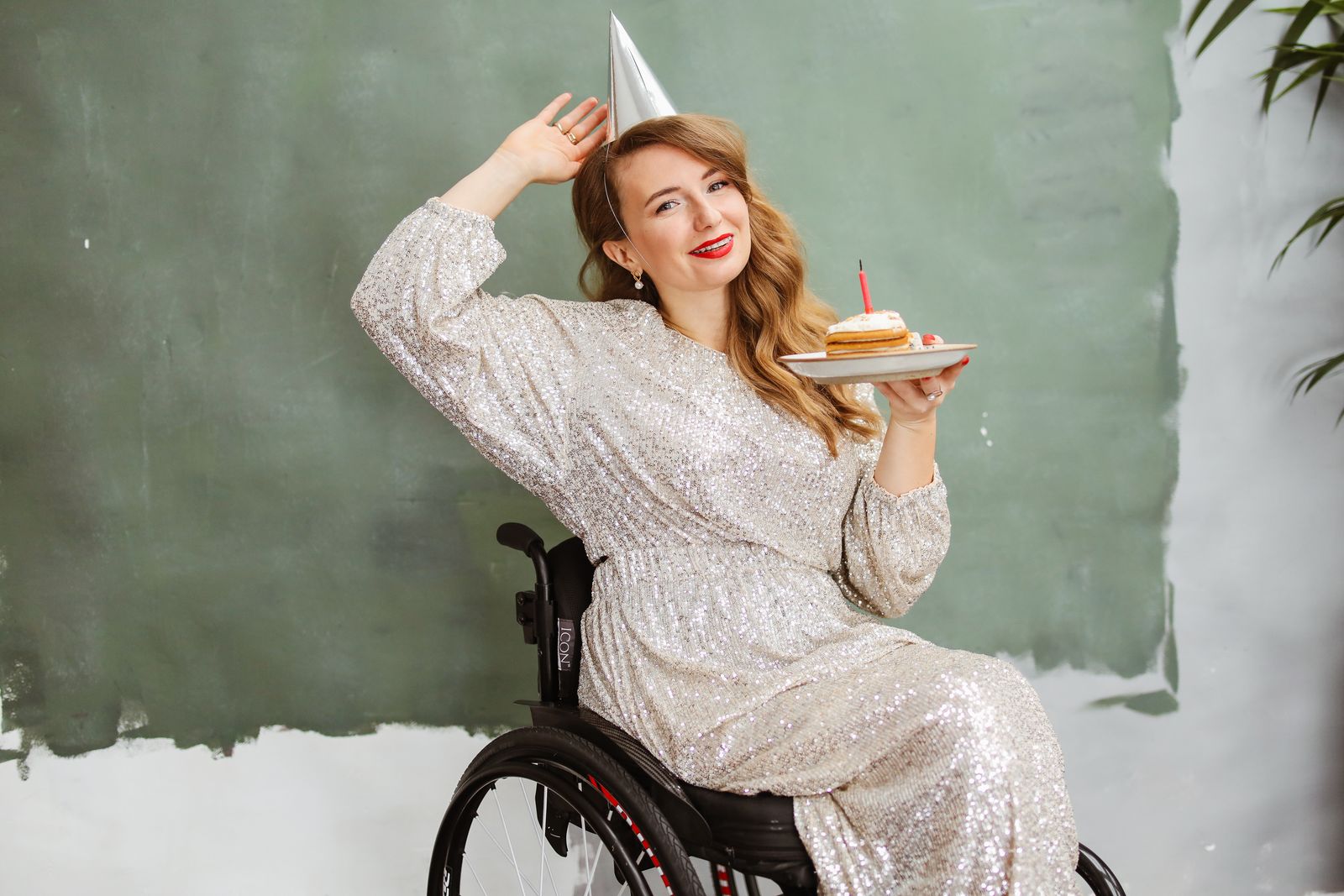 A smiling wheelchair user wearing a party hat and holding a birthday cake