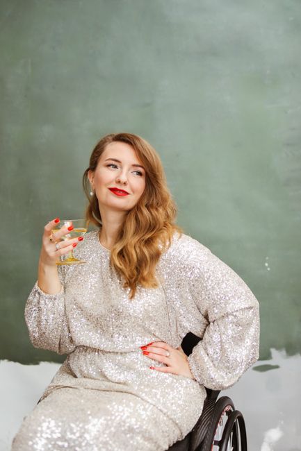 Caucasian woman in a sequined dress seated in a wheelchair holding a wine glass, exuding elegance and glamour.