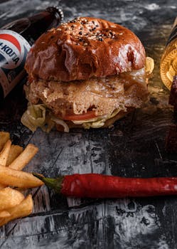 From above appetizing beef burger and french fries placed on gray table near fizzy drink bottle and red chili pepper