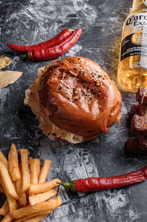 From above composition of yummy beef burger placed on gray table near yummy french fries red chili peppers and cold beer