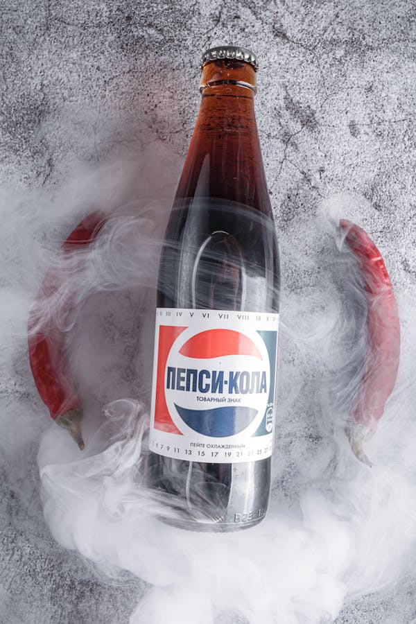 Top view composition of glass bottle of nonalcoholic cold refreshing soft drink placed on gray table near red chili peppers and covered with crawling dry ice smoke