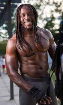 Smiling shirtless man with braided hair working out outdoors with resistance bands.