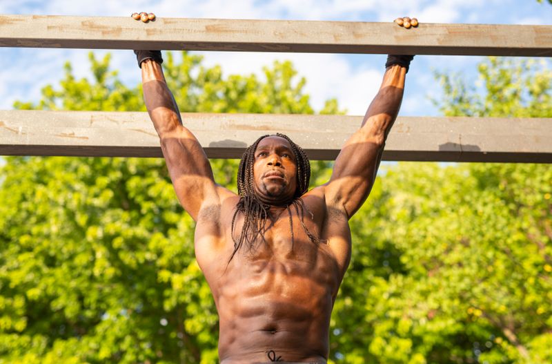Shirtless African American man performing pull-ups in outdoor park setting.