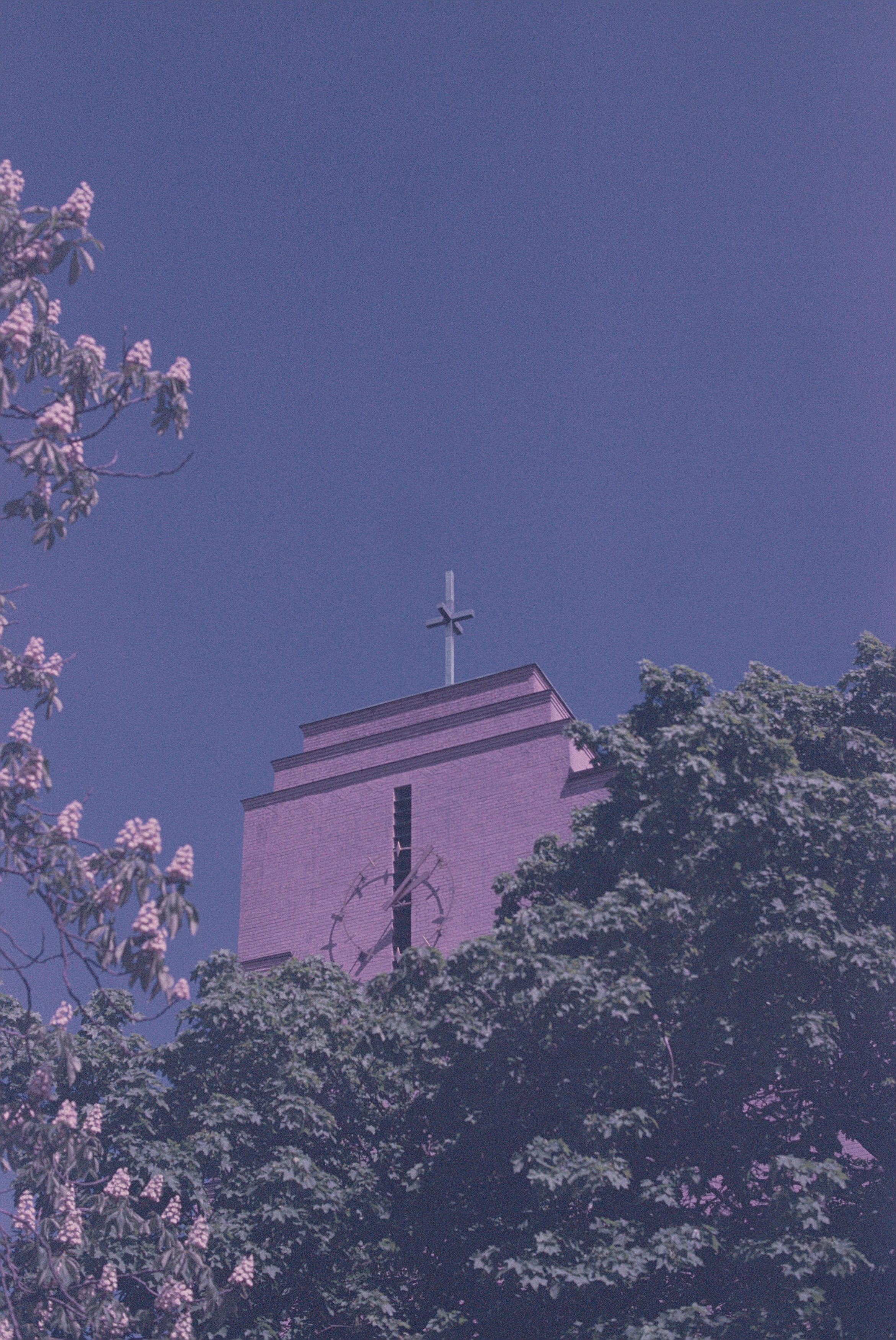 A tall church tower with a cross visible above lush trees under a clear sky.