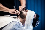 An adult man receiving a precise beard trim in a barbershop with a warm towel treatment.