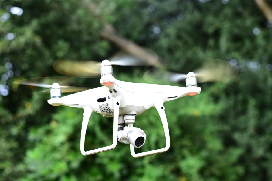 White drone hovering outdoors with blurred green trees in background.