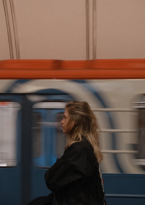 Side view contemplating young female in black jacket standing on underground station platform near departing train and looking away thoughtfully